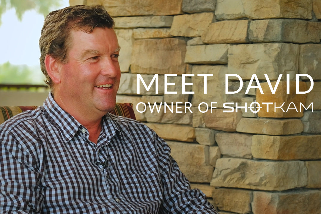 David Stewart, owner of ShotKam, sitting outside in front of a stone wall, smiling during an interview, wearing a checkered shirt.