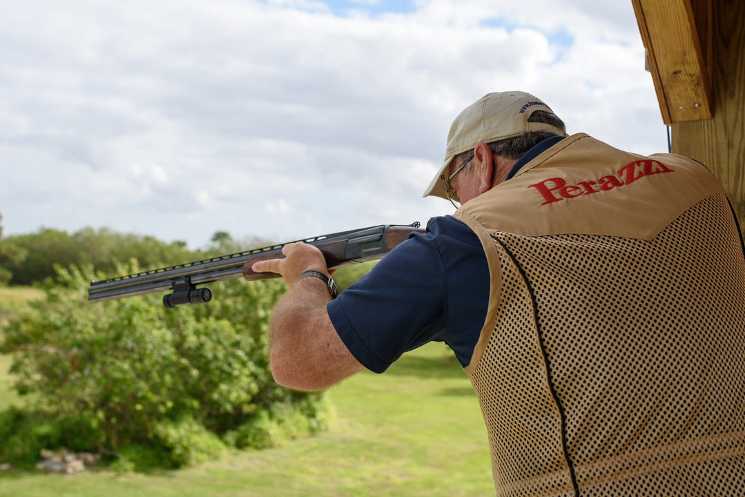 Back view of a shooter in a Perazzi vest aiming a shotgun from a shooting stand, focused on the target in an outdoor clay shooting range.