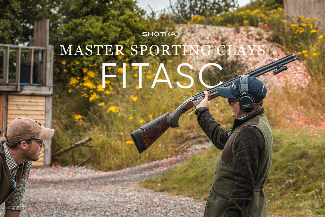 Shooter lifting over-under shotgun at a FITASC course with clays scattered on the ground, ShotKam mounted on barrel.