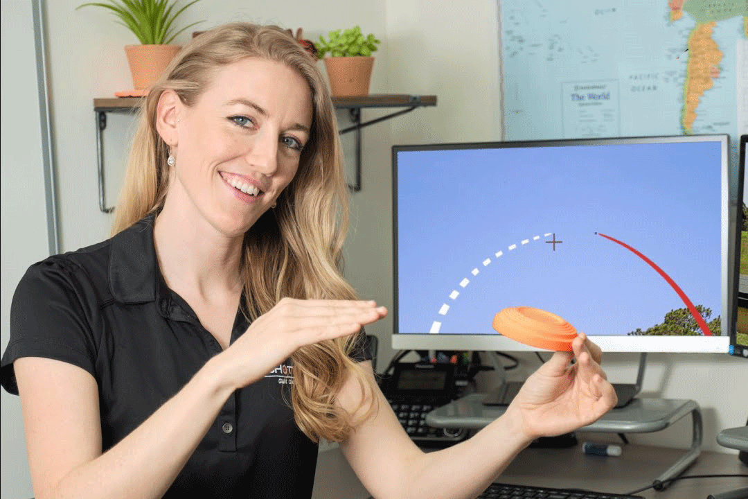 Emily Stewart, COO of ShotKam, is demonstrating a shooting concept using an orange clay target in her hands. Behind her is a computer screen displaying a trajectory of a clay target with dotted lines and a red arc, representing a shot path.