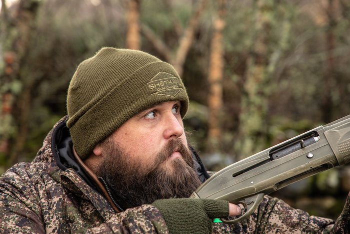 Bearded hunter in camouflage gear and olive-green ShotKam beanie, focusing intently while holding a shotgun, surrounded by a woodland backdrop.