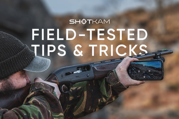 Hunter aiming a shotgun with a ShotKam camera mounted on the barrel while reviewing footage on a smartphone in the field.