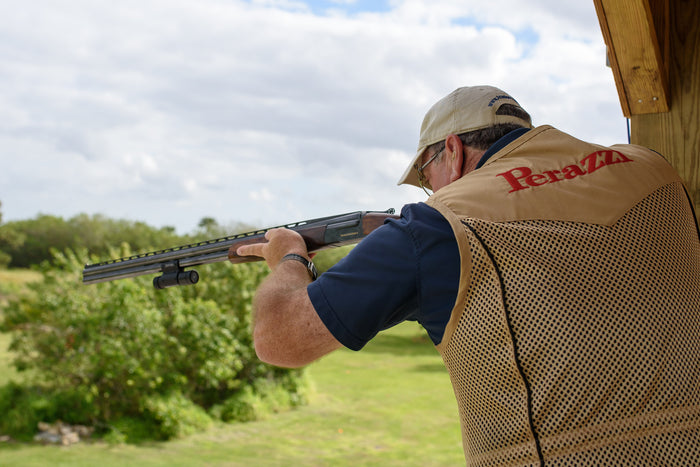 Back view of a shooter in a Perazzi vest aiming a shotgun from a shooting stand, focused on the target in an outdoor clay shooting range.