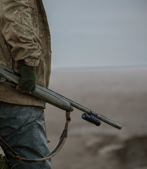 Hunter holding a shotgun equipped with a ShotKam Gun Camera, dressed in camouflage gear and gloves, standing outdoors in a rugged environment. The ShotKam is clearly visible mounted on the shotgun barrel, ideal for recording hunting adventures and shooting accuracy. This image captures the essence of hunting, emphasizing the utility of ShotKam products for capturing every moment. 