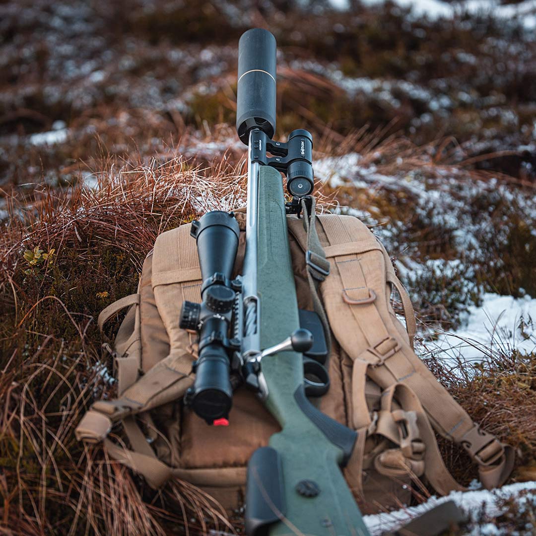 Bolt-action rifle equipped with the ShotKam Gen 4 using the Moderator Mount, resting on a tactical backpack in a rugged outdoor setting. Perfect for American hunters and precision shooters looking to record high-definition footage of their shots.