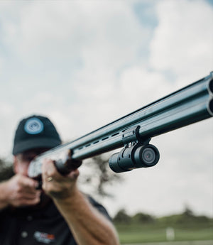 of ShotKam owner and David aiming a shotgun equipped with a ShotKam Gun capturing every moment of the shooting The ShotKam is mounted securely on the shotgun ideal for recording target practice and hunting The shooter wears a cap and focusing intently on the target against a backdrop of a partly cloudy